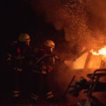 Two firefighters fighting a heavy car fire with water, 2 men in black helmet standing near fire